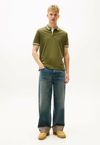 Oliwkowa koszulka polo z białym i czarnym paskowanym kołnierzykiem, noszona z szerokimi niebieskimi dżinsami i brązowym obuwiem z teksturowaną podeszwą.