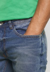 Persona vestida con jeans de mezclilla azul y una camiseta verde, con la mano derecha metida en el bolsillo delantero de los jeans.