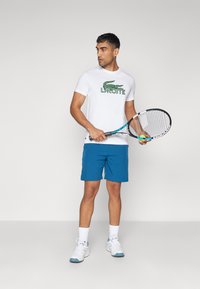 Camiseta blanca de manga corta con el logo verde de Lacoste, combinada con pantalones cortos azules y zapatillas deportivas blancas, sosteniendo una raqueta de tenis.