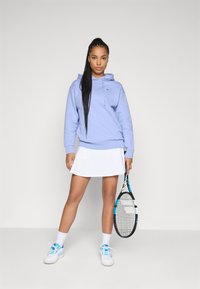 Hanorac violet deschis cu sireturi, fustă de tenis albă cu perforații, adidași albi cu accente albastre, ținând o rachetă de tenis.