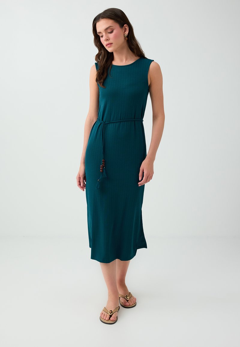 Femme aux longs cheveux foncés portant une robe midi turquoise sans manches avec une fine ceinture et des sandales marron, debout devant un fond clair uni.