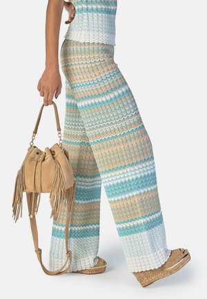 Femeie purtând pantaloni largi cu dungi multicolore din tricot și un top asortat, ținând o geantă bej din piele intoarsă cu franjuri, purtând sandale cu platformă.