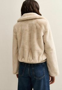Chaqueta de piel sintética beige con cuello, dobladillo elástico y mangas largas, combinada con vaqueros oscuros de cintura alta.