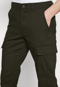 Ciemnozielone spodnie cargo z boczną kieszenią, zapięciem na guzik i dopasowanym krojem. Materiał wydaje się wytrzymały o gładkiej fakturze.