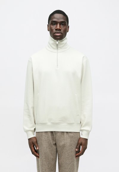 Mann in einem weißen, hoch geschlossenen Reißverschluss-Sweatshirt und braunen strukturierten Hose, der vor einem schlichten hellen Hintergrund steht.