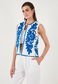 White sleeveless vest with blue floral embroidery and trim, featuring a mid-front opening. Worn over a white top and paired with light trousers.