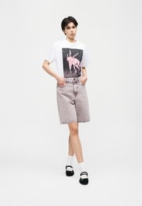 Personne debout, les mains dans les poches, portant un t-shirt blanc avec un motif d’âne rose, un short en jean clair, des chaussettes blanches et des chaussures noires.