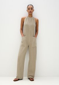 Beige Jumpsuit aus leichtem Stoff mit Halterneck, vorderem Knopfverschluss, zwei großen Taschen und weitem Bein. Strukturierte Oberfläche.