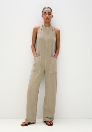 Beige jumpsuit made of lightweight fabric with a halter neck, front button closure, two large pockets, and wide legs. Textured finish.