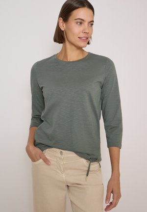 Femme aux cheveux courts et bruns portant un haut gris à manches longues avec des cordons et un pantalon beige, regardant vers la droite avec une main dans la poche.