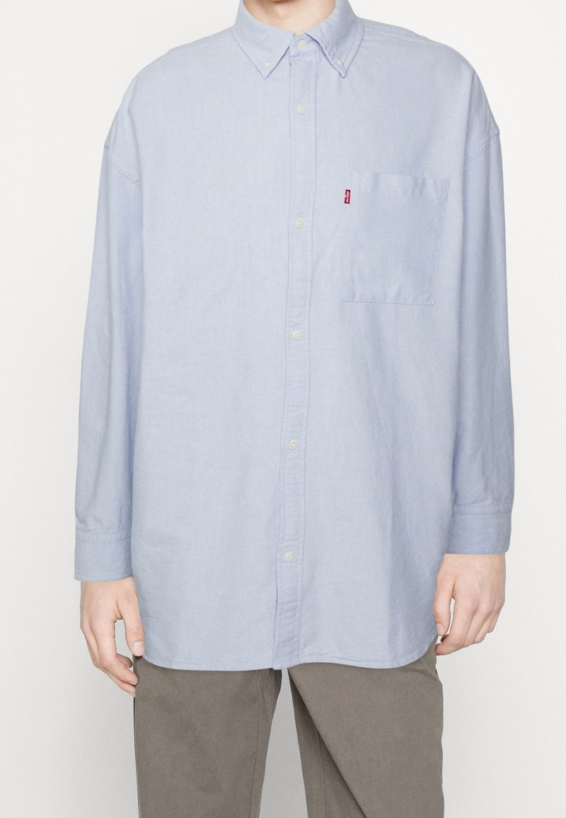 Henkilö, jolla on yllä vaaleansiniset pitkät hihat napitettava paita rintataskulla ja ruskeat housut, seisoo tasaisella taustalla.