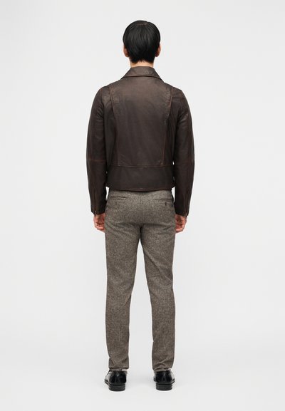 Άνδρας με σκούρο καφέ δερμάτινο μπουφάν, γκρίζο υφασμάτινο παντελόνι και μαύρα παπούτσια, στέκεται με την πλάτη προς το μέρος μας σε απλό ανοιχτό φόντο.