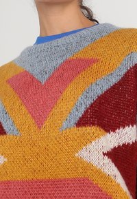 Pull en tricot avec un motif géométrique en marron, jaune, rose et bleu clair, doté d'un col rond et d'un tissu texturé.