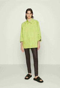 Camisa oversized verde lima, abotoada com colarinho, combinada com calças escuras ajustadas e sandálias pretas de deslizar. Tecido liso com textura mínima.