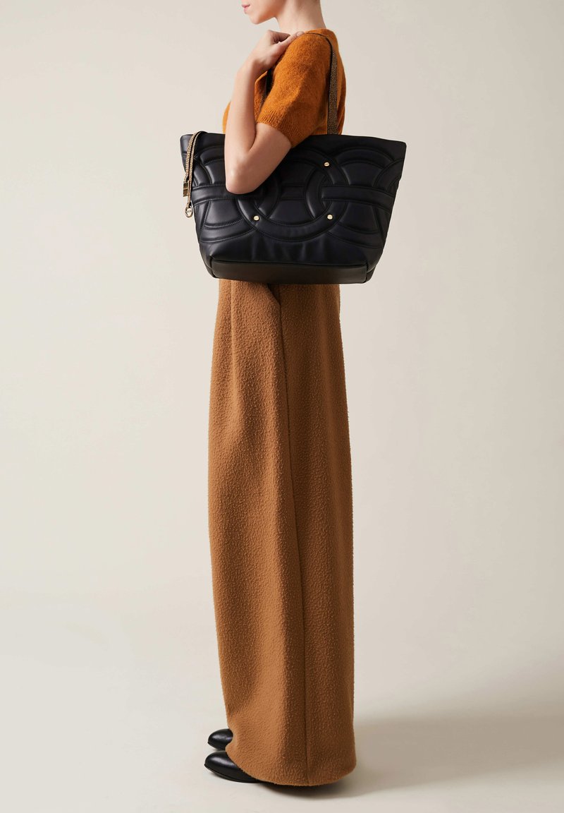 Borsa tote in pelle nera con motivo testurizzato, dettagli dorati e cerniera. Il modello indossa un completo in misto lana marrone con scarpe nere.