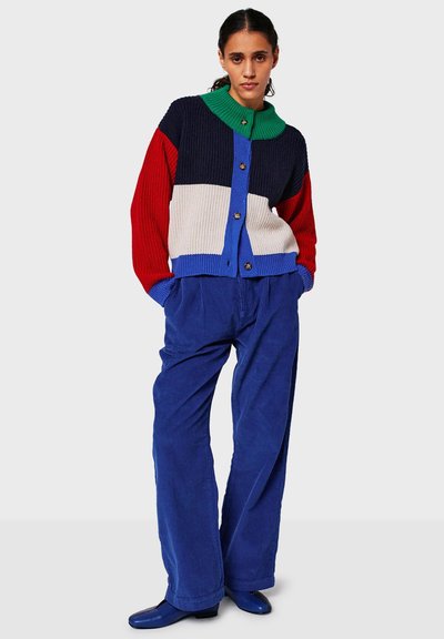 Monivärinen neulecardigan, jossa on punaisia, sinisiä, vihreitä ja kermaisia osioita, napitettava etuosa ja ribbitekstuuri. Yhdistetty sinisiin corduroy-housuihin.