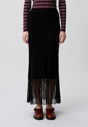 Donna che indossa una gonna midi plissettata nera con orlo trasparente, top a maniche lunghe a righe color bordeaux e scarpe chiuse in pelle marrone.