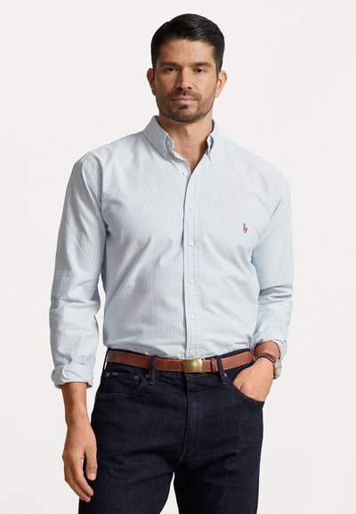 Hellblau-weiß gestreifte Hemd mit Knopfleiste und klassischem Kragen, getragen mit dunklen Jeans und einem braunen Ledergürtel. Gesticktes Logo auf der Brust.