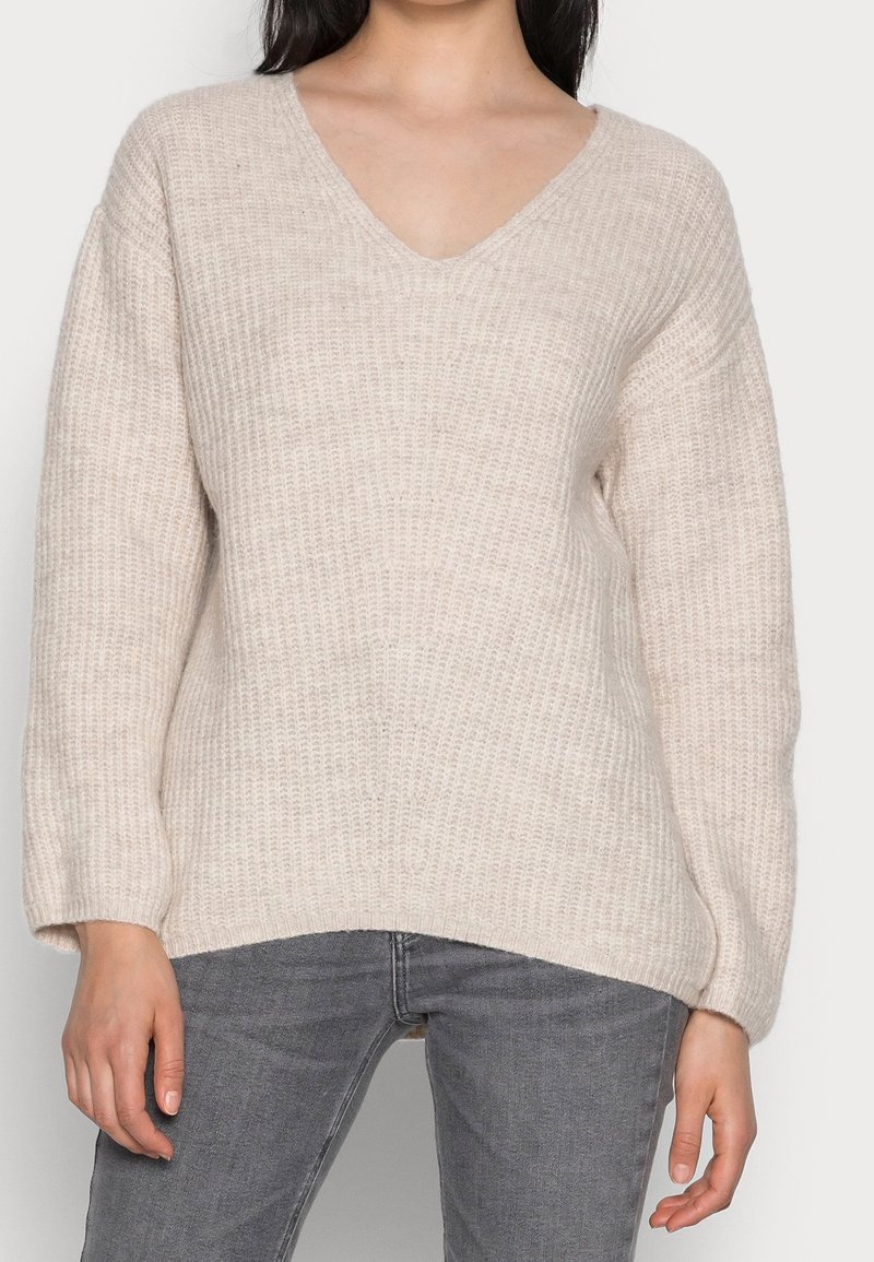 Vrouw in een beige gebreide V-hals trui met lange mouwen en een grijze spijkerbroek, staande voor een effen witte achtergrond.