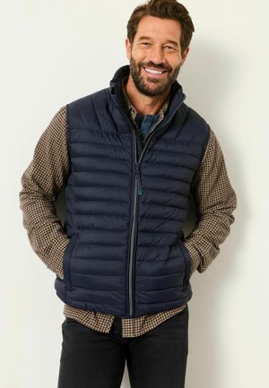Lächelnder Mann trägt eine marineblaue Steppweste über einem braun karierten Hemd, die Hände in den Taschen und steht vor einer einfarbigen hellen Wand.