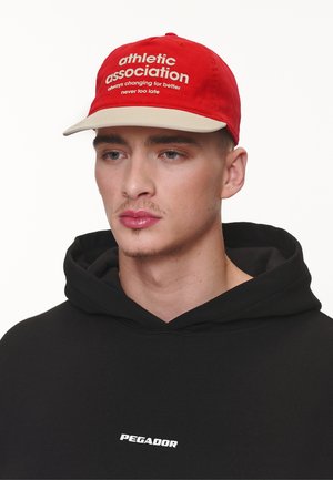 Casquette en coton rouge avec un bord beige, portant un texte blanc qui dit "association athlétique" et "toujours en évolution pour le mieux."