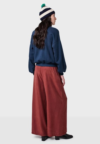 Navyblaues Sweatshirt mit überschnittenen Schultern, kombiniert mit weit geschnittenen, rostfarbenen Hosen. Das Modell trägt eine gestreifte Strickmütze mit einem Pompon.