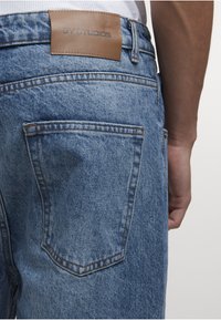 Niebieskie jeansy z denimu z tylną kieszenią i brązową skórzaną metką z napisem "2Y STUDIOS", noszone przez osobę z widocznym ramieniem u boku.