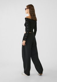 Czarny, prążkowany top typu off-the-shoulder z ozdobnymi guzikami na rękawach, zestawiony z luźnymi czarnymi spodniami i srebrnymi sneakersami. Gładka faktura.