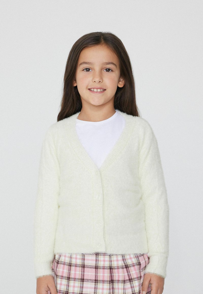 Flauschige, hellgelbe Strickjacke mit V-Ausschnitt und Knopfverschluss, die lange Ärmel und eine weiche Textur hat, kombiniert mit einem karierten Rock.