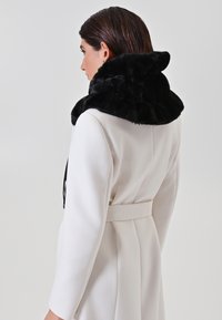 Cappotto bianco con vita cintata, caratterizzato da un morbido colletto in pelliccia sintetica nera. Texture liscia con un design strutturato, dettagli di cuciture visibili.