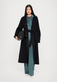 Garš melns mētelis ar jostu, uzvilkts virs zila kordūrija kostīma. Modelis tur melnu ādas somu; baltas papēžu kurpes papildina apģērbu.