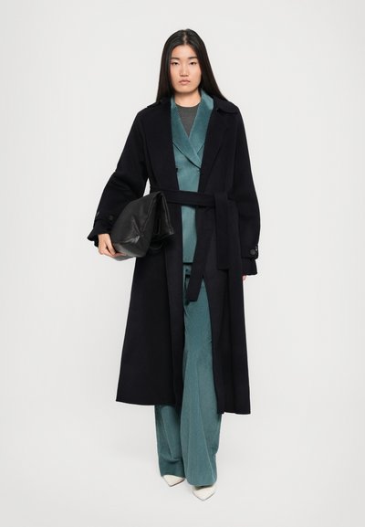 Largo abrigo negro con un cinturón, llevado sobre un traje de pana en color teal. Modelo sosteniendo un bolso de cuero negro; zapatos de tacón blancos completan el atuendo.