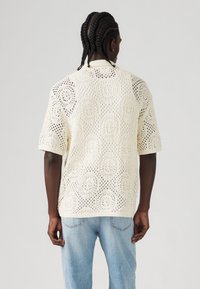 Man met cornrow vlechten in een crèmekleurig gehaakt kortemouwen trui en lichtblauwe jeans, met zijn rug naar de camera gericht op een effen achtergrond.