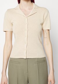 Polo shirt lavorato a maglia beige a maniche corte con colletto, dotato di una chiusura con bottoni e un design aderente. Texture liscia con dettagli a costine.