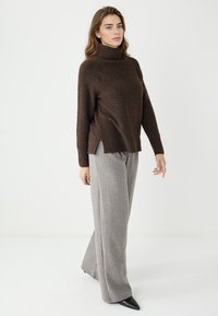 Brauner Strickrollkragenpullover mit gerippten Manschetten und seitlichen Schlitzen, kombiniert mit grauen weiten Hosen und schwarzen spitzen Schuhen.