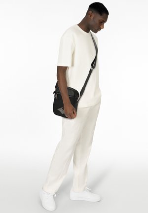 Borsa a tracolla nera con zip, tracolla regolabile e superficie texturizzata. Il modello indossa una t-shirt crema e pantaloni chiari con sneaker bianche.