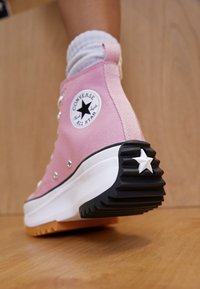 Pink Converse high-top sneaker with a black and white star logo, white eyelets, textured black outsole, and orange bottom accent.