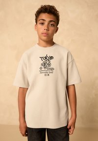 T-shirt beige chiaro a maniche corte con motivo a griglia testurizzata e stampa grafica nera raffigurante una tigre e la parola "TOKYO". Indossata con pantaloni scuri.
