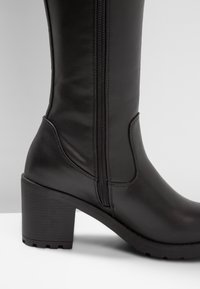 Czarny skórzany bucik do połowy łydki z bocznym zamkiem i słupkowym obcasem, przedstawiony na jednolitym szarym i białym tle.