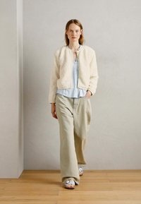 Casaco bomber texturizado off-white sobre uma blusa azul clara, combinado com calças largas bege e sandálias brancas em um chão de madeira.