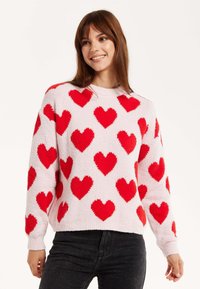 Fuzzy pink sweater featuring large red hearts in a scattered pattern, ribbed neckline, and cuffs, paired with dark jeans.