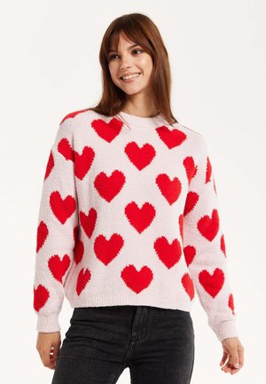 Fuzzy pink sweater featuring large red hearts in a scattered pattern, ribbed neckline, and cuffs, paired with dark jeans.