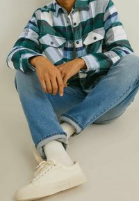 Koszula flanelowa w kratę w kolorach teal, biały i niebieski; lekkie dżinsy; kremowe buty; siedząca poza, ręce spoczywają na kolanach, swobodny strój.