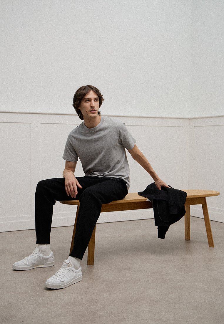 Grijze t-shirt, zwarte broek en witte sneakers. Het onderwerp zit op een houten bank in een minimalistische, lichtgekleurde kamer.