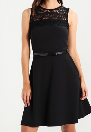 Černé šaty bez rukávů s krajkovým překrytím u výstřihu, přiléhavým pasem s tenkým páskem a rozšířenou sukní, které nosí stojící žena.