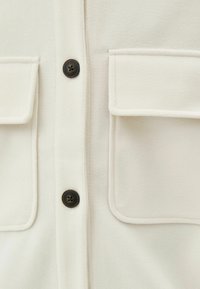 Camicia button-up color crema con due tasche a busta, caratterizzata da bottoni scuri e una texture morbida. Design minimalista con linee pulite.