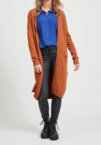 Kopparfärgad lång kardigan i mjukt stickat material, med öppen front, ribbade manschetter och två sidofickor, kombinerad med en blå skjorta och svarta byxor.