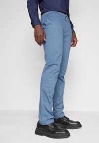 Pantaloni azzurri chiari con una texture liscia, taglio dritto e due tasche frontali, abbinati a scarpe nere di pelle con suola spessa.