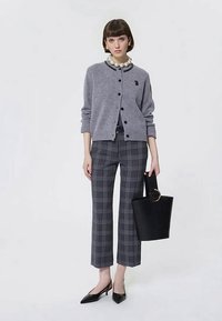 Jeune femme portant un cardigan gris boutonné, un pantalon écossais court, des talons aiguilles noirs, un collier de perles, et tenant un sac à main noir.