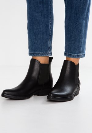 Bottes chelsea en cuir noir avec panneaux élastiques sur les côtés, texture lisse, bout arrondi et petits talons empilés. Portées avec un jean bleu raccourci.
